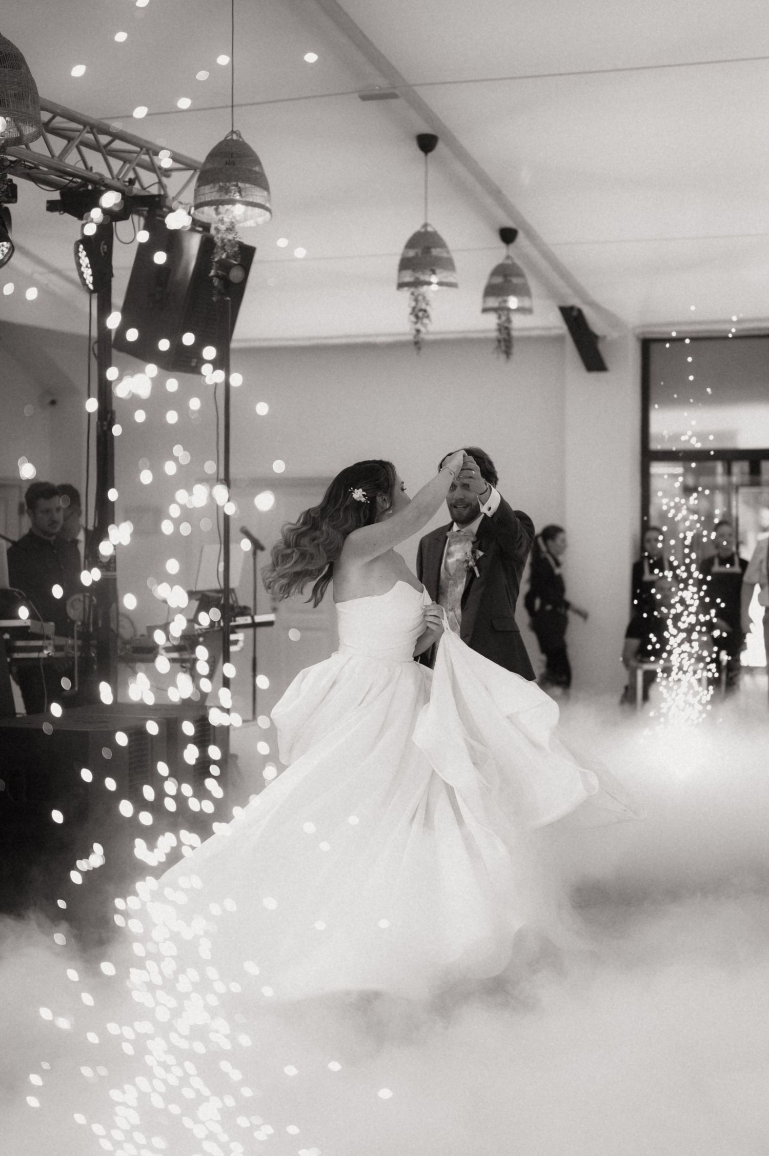 Black and white photo of bride and groom sharing first dance surrounded by sparkling fireworks and dreamy fog on wedding day.