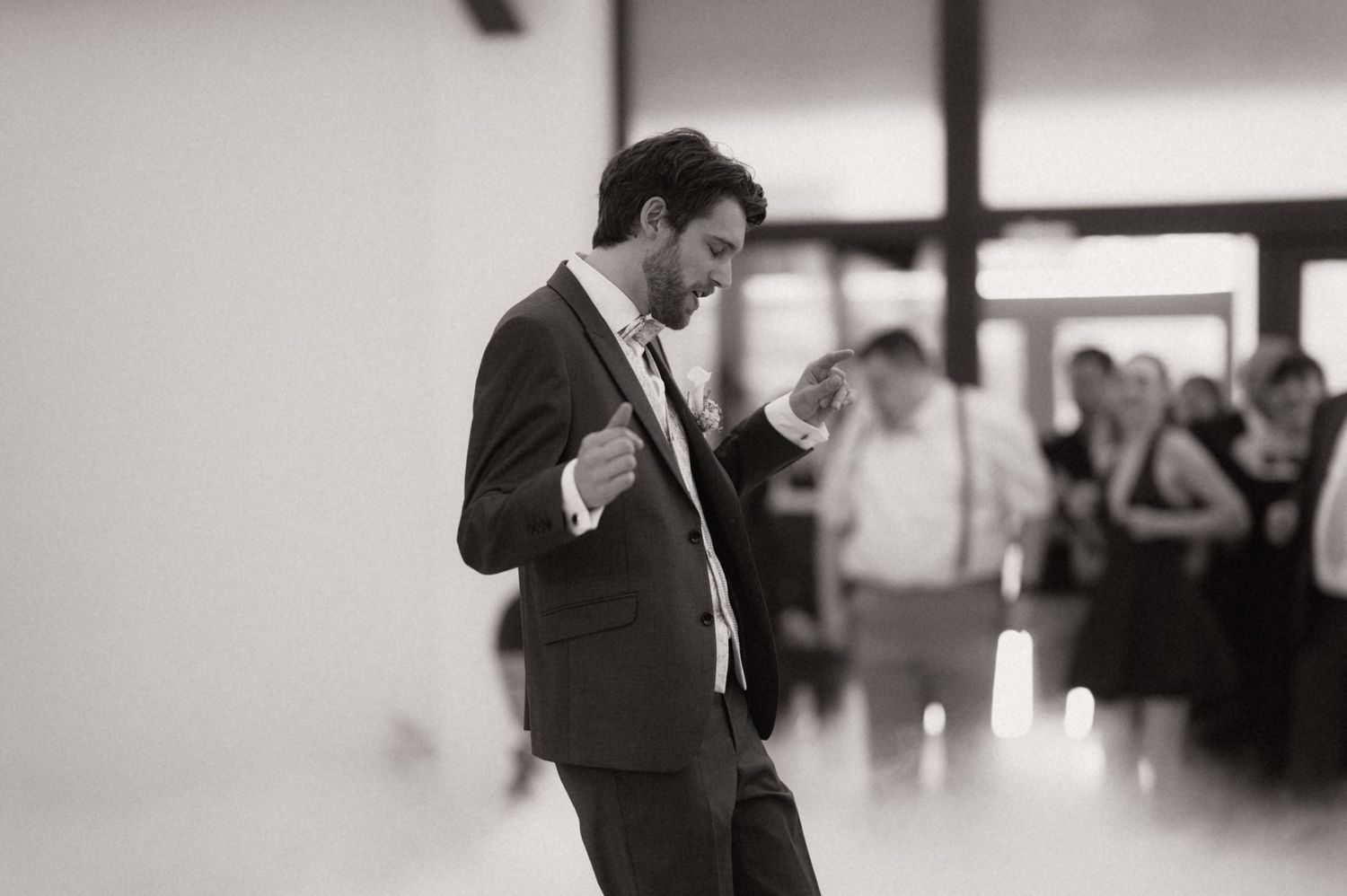Man in suit dancing energetically at a wedding reception, black and white photo with guests in background.