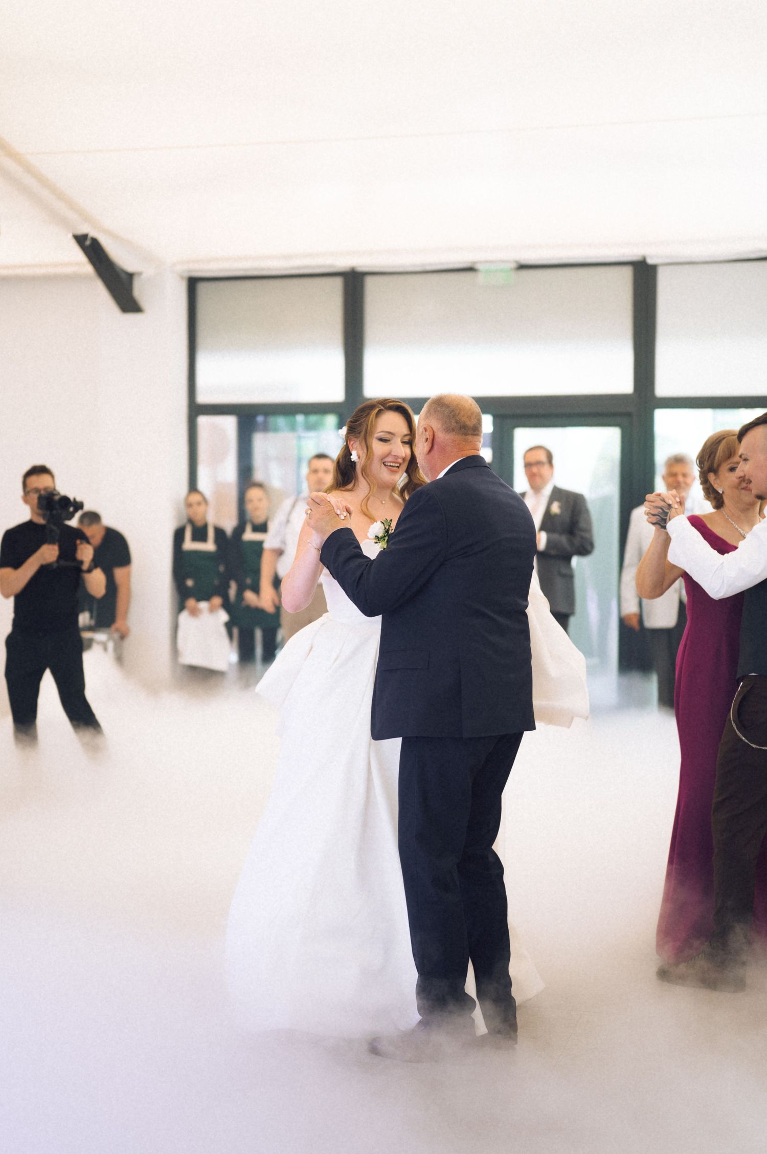 Bride and groom share first dance on fog-covered floor at elegant wedding reception with guests watching.