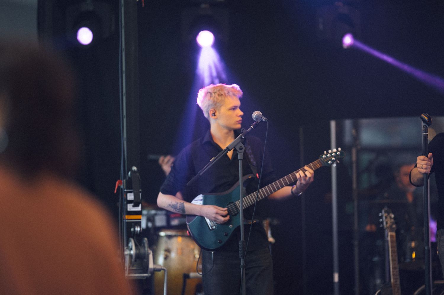 Blonde guitarist performing on stage under purple lights at a live music concert event.