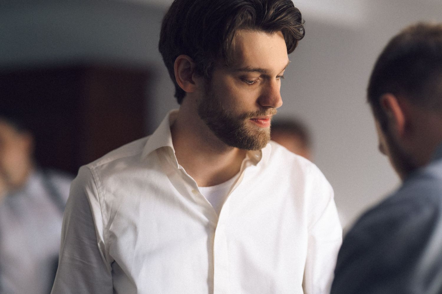Young bearded man in white shirt looking down thoughtfully in a softly lit indoor setting.
