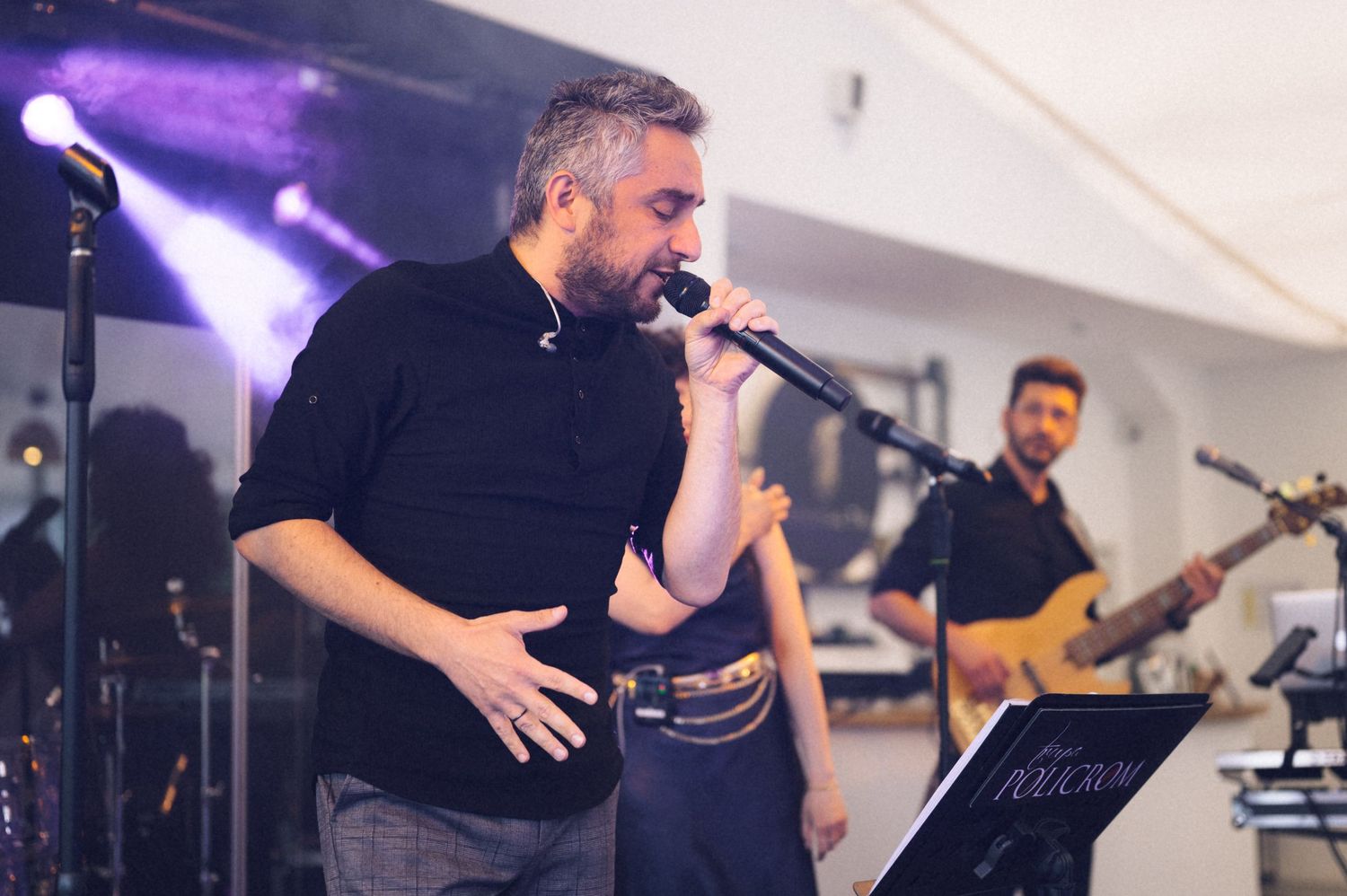 Male singer performing on stage with microphone, band member playing bass guitar in background at live event.