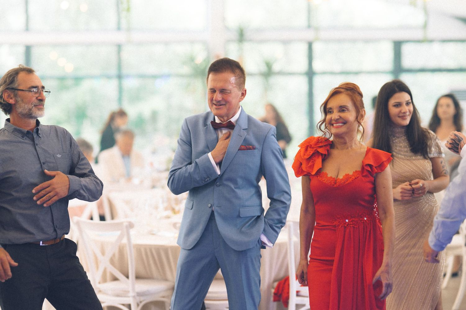 Guests in formal attire laugh and dance at an elegant wedding reception in a bright venue.