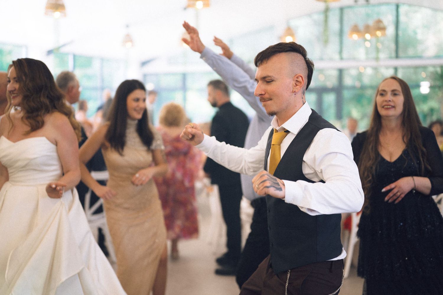 Guests dancing joyfully at a wedding reception, with groom in vest and gold tie celebrating on the dance floor.