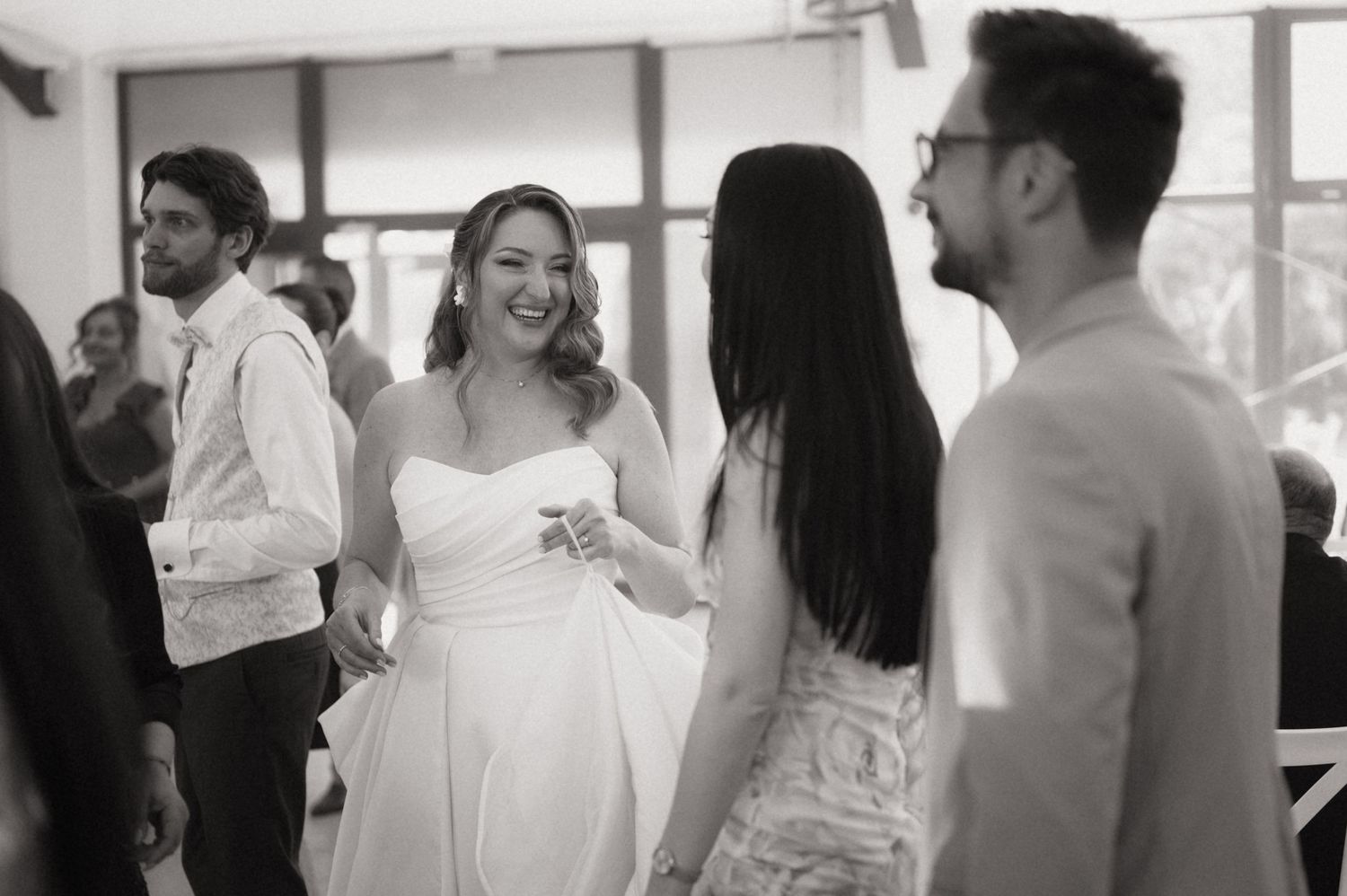 Black and white photo of a smiling bride in a strapless gown chatting with guests at a wedding reception.