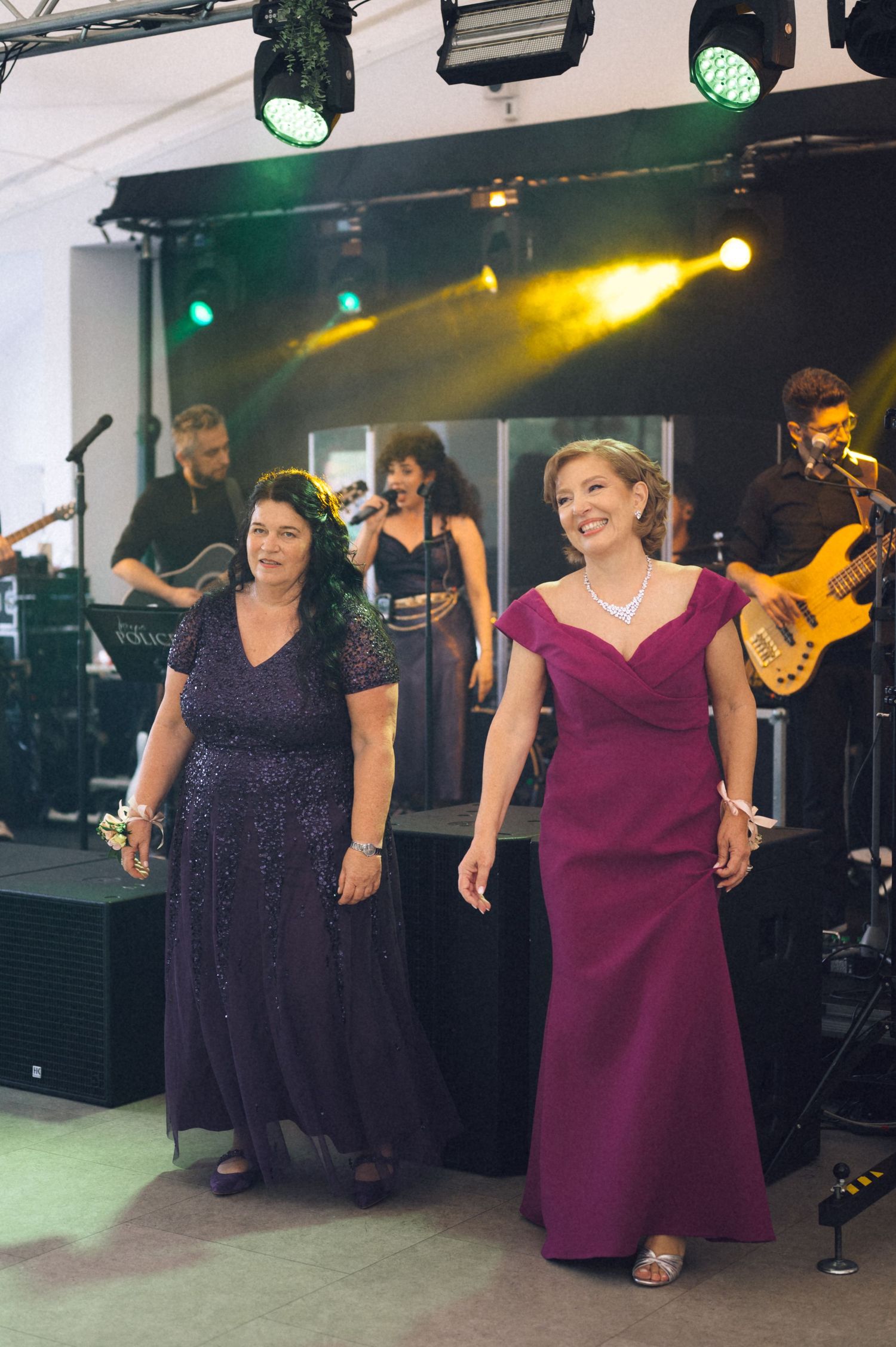 Two women in elegant purple gowns walk at a wedding reception with a live band performing on stage behind them.