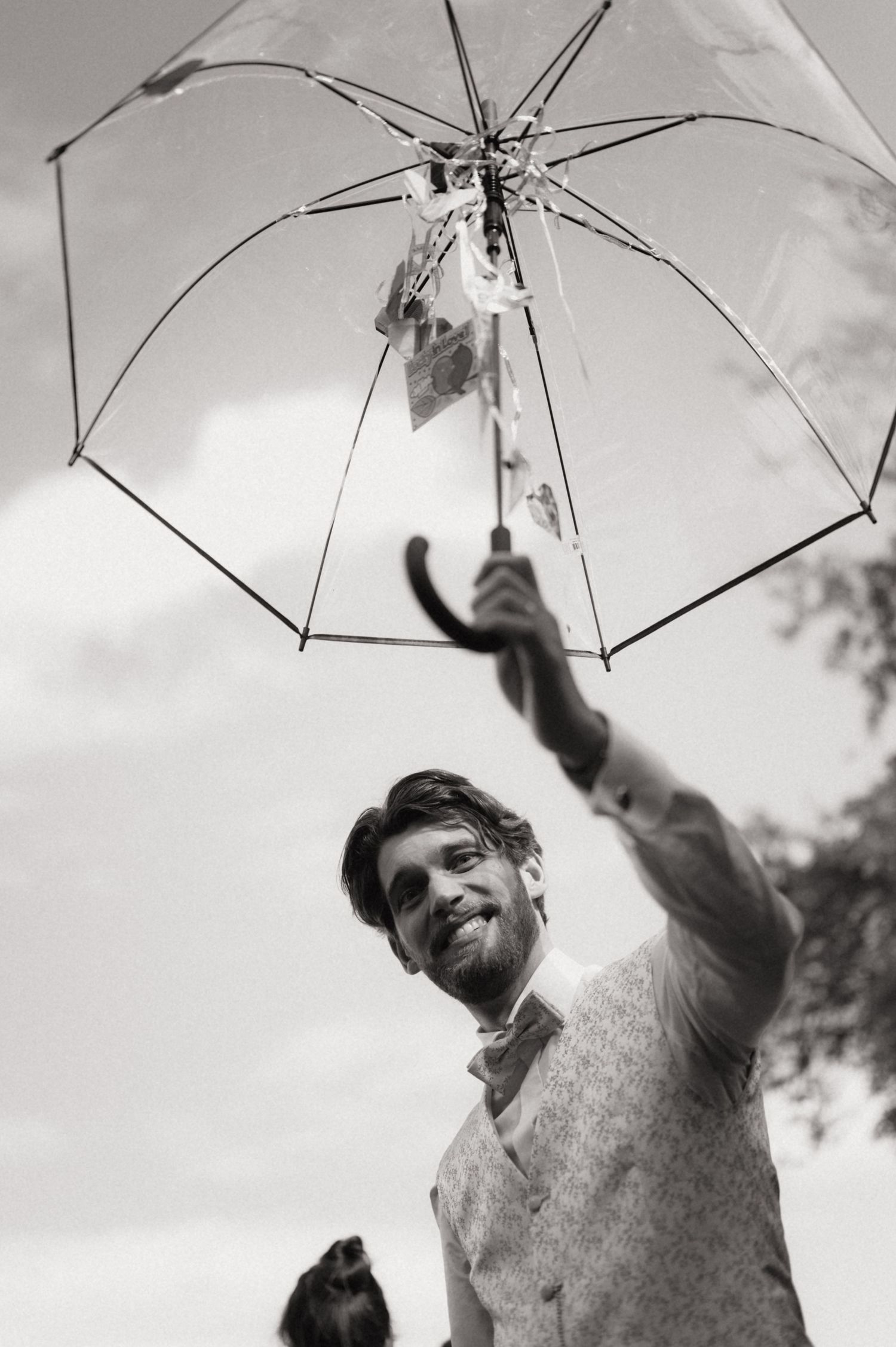 Black and white photo of a smiling bearded man holding a transparent umbrella overhead, shot from below against a cloudy sky.
