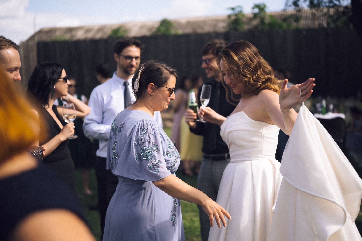 Bride in white gown chatting with guest in blue dress at outdoor wedding reception, guests mingling in background.