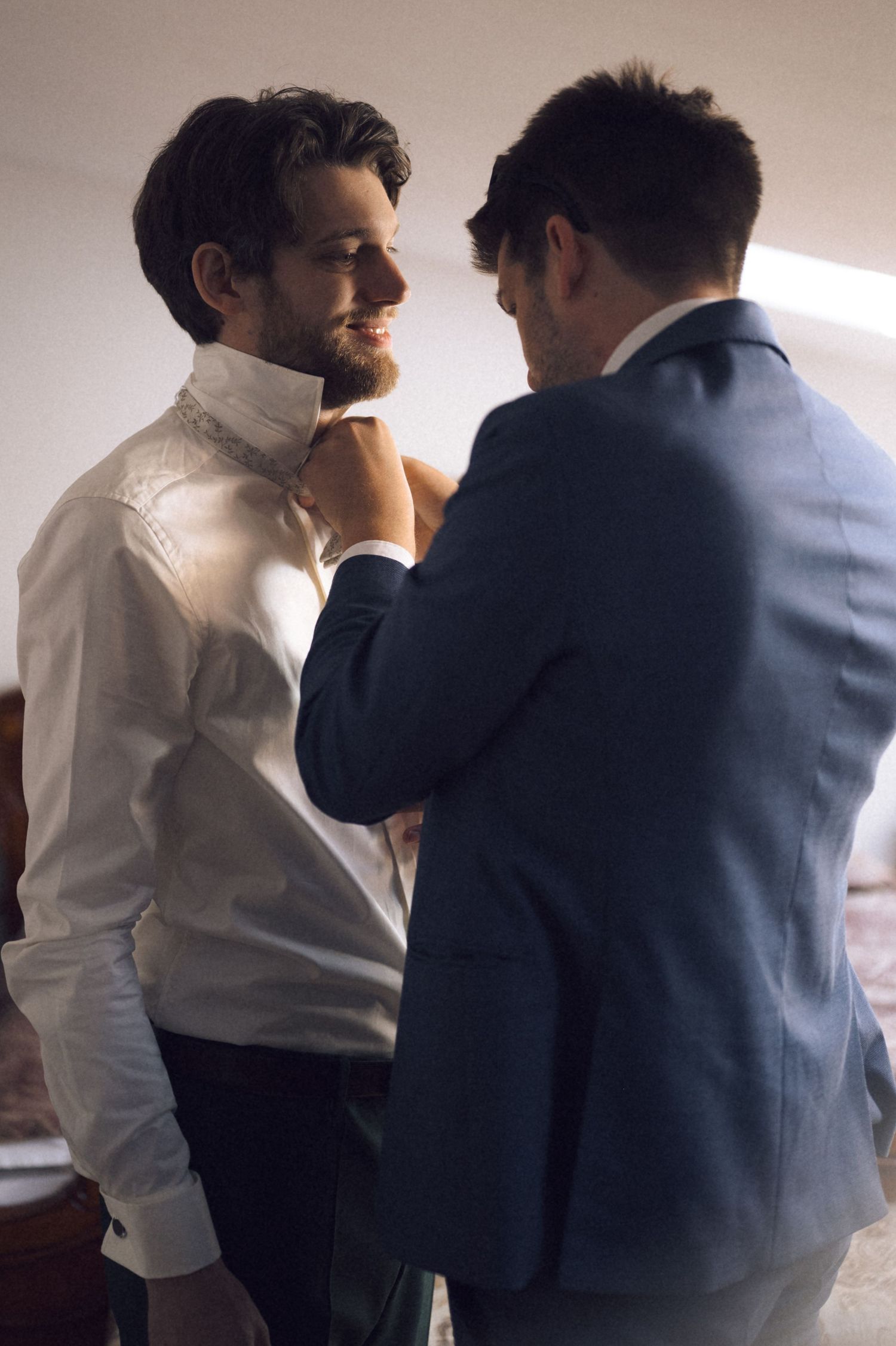 Two well-dressed men in suits adjusting a tie, preparing for a formal event or wedding ceremony.