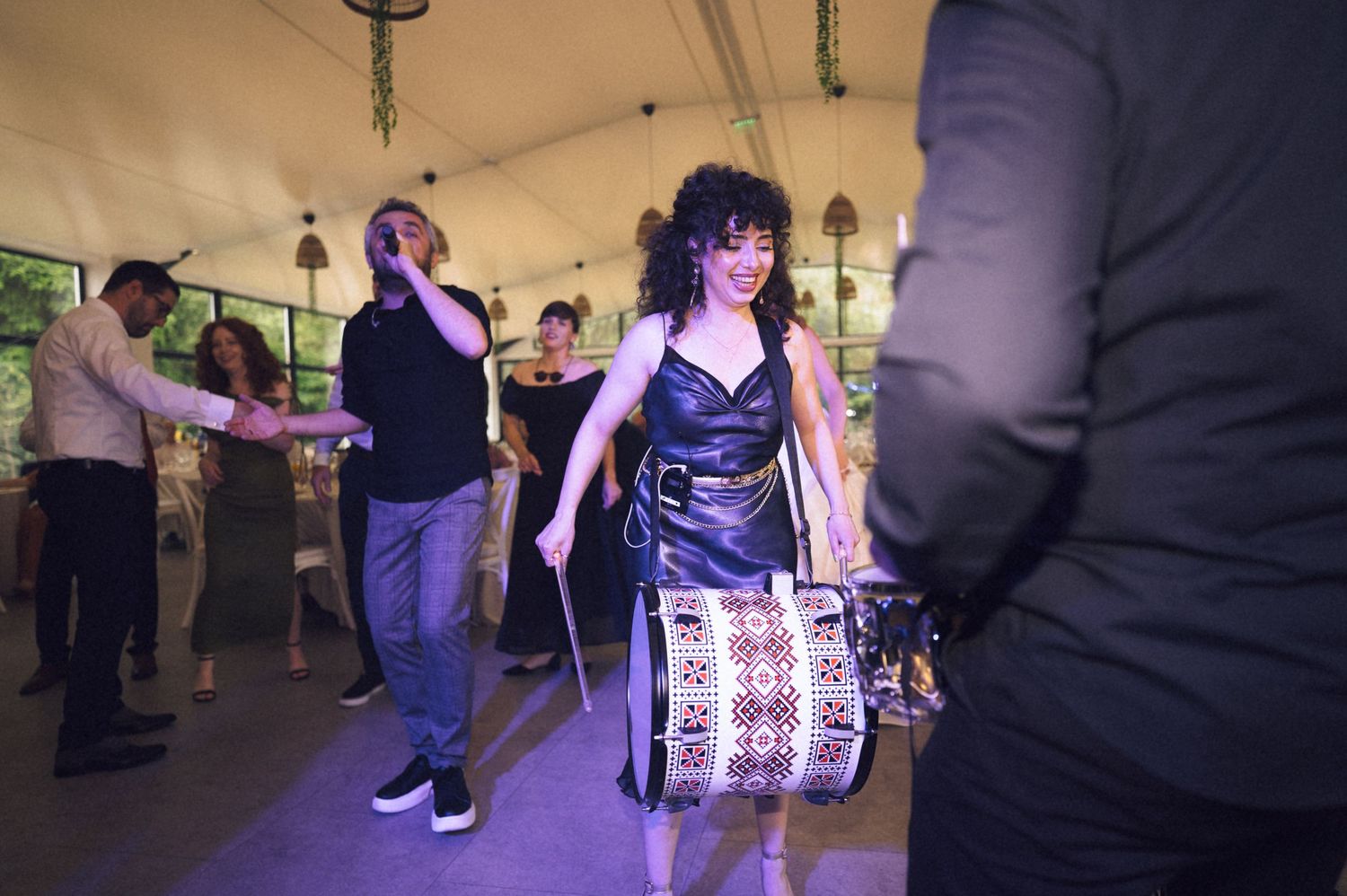 Woman in black dress playing a decorative drum at a lively indoor party with guests dancing under purple lights.