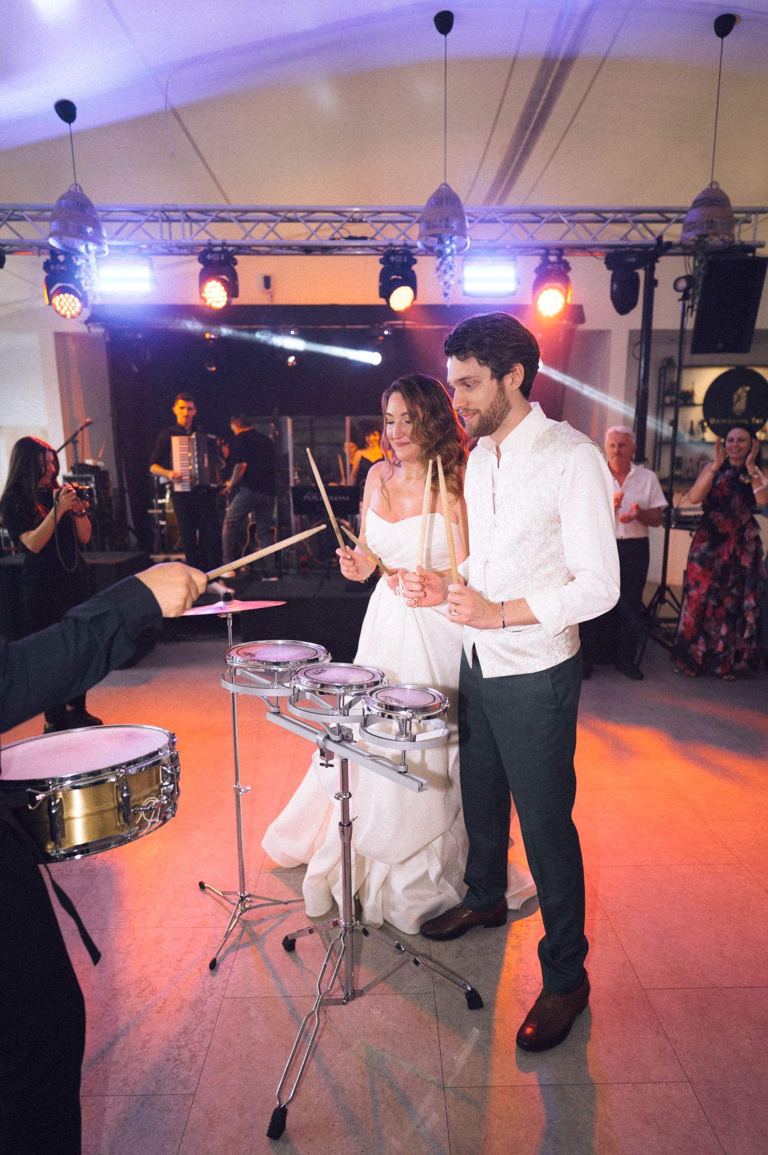 Bride and groom playing drums together at wedding reception with live band performing on stage behind them.