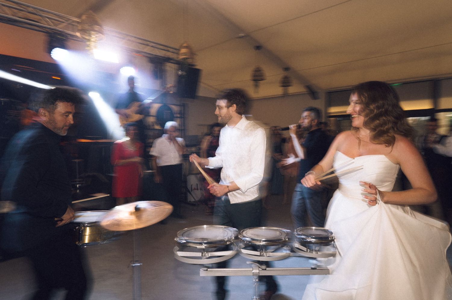 Bride in white gown and groom in white shirt playing drums together at a lively wedding reception party.
