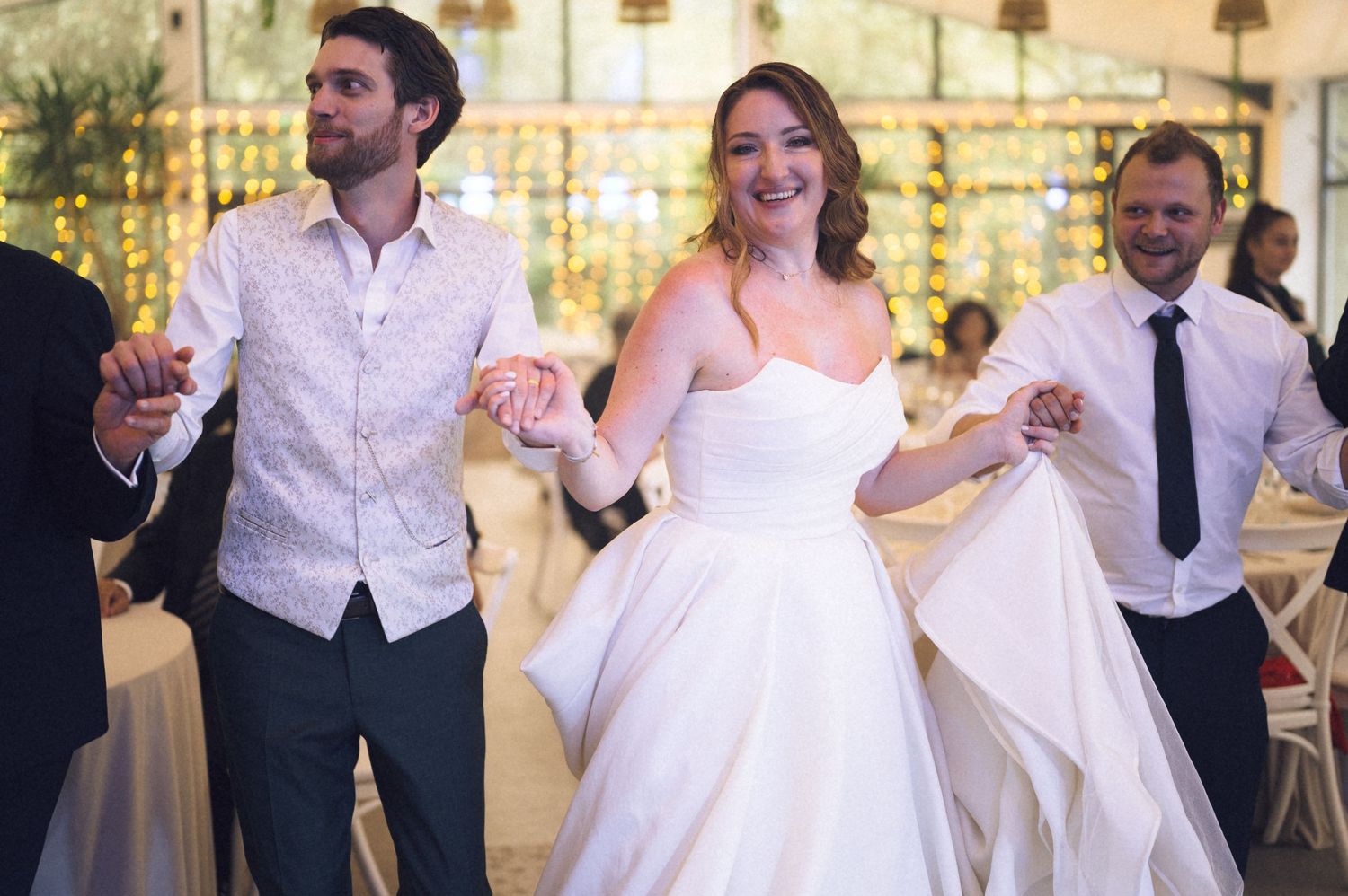 Bride in white strapless gown dancing joyfully with groom in vest and guest at elegant wedding reception.