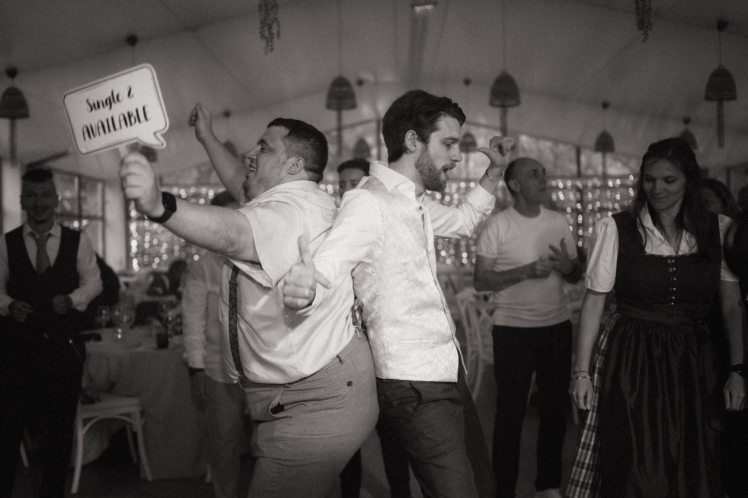 Two men dancing joyfully at a wedding reception, one holding a humorous sign, captured in black and white.
