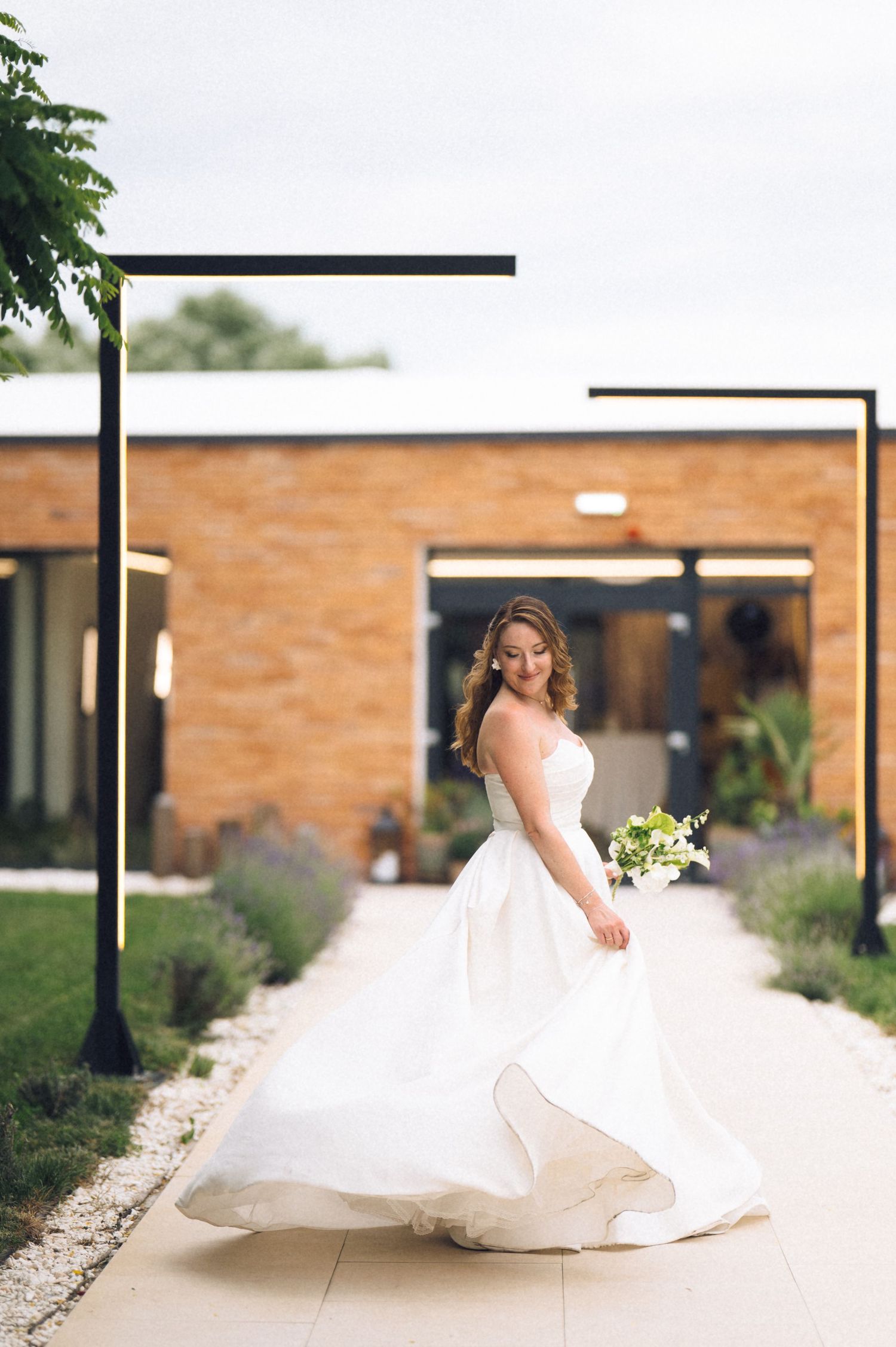 Bride in a flowing white ballgown twirling on a garden path, holding a green bouquet, with a modern brick building backdrop.