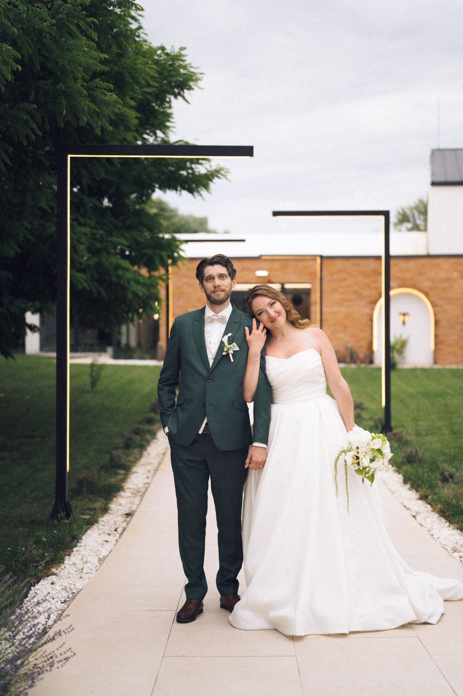 Bride and groom posing under a modern geometric arch on a garden pathway, bride in white gown holding bouquet.