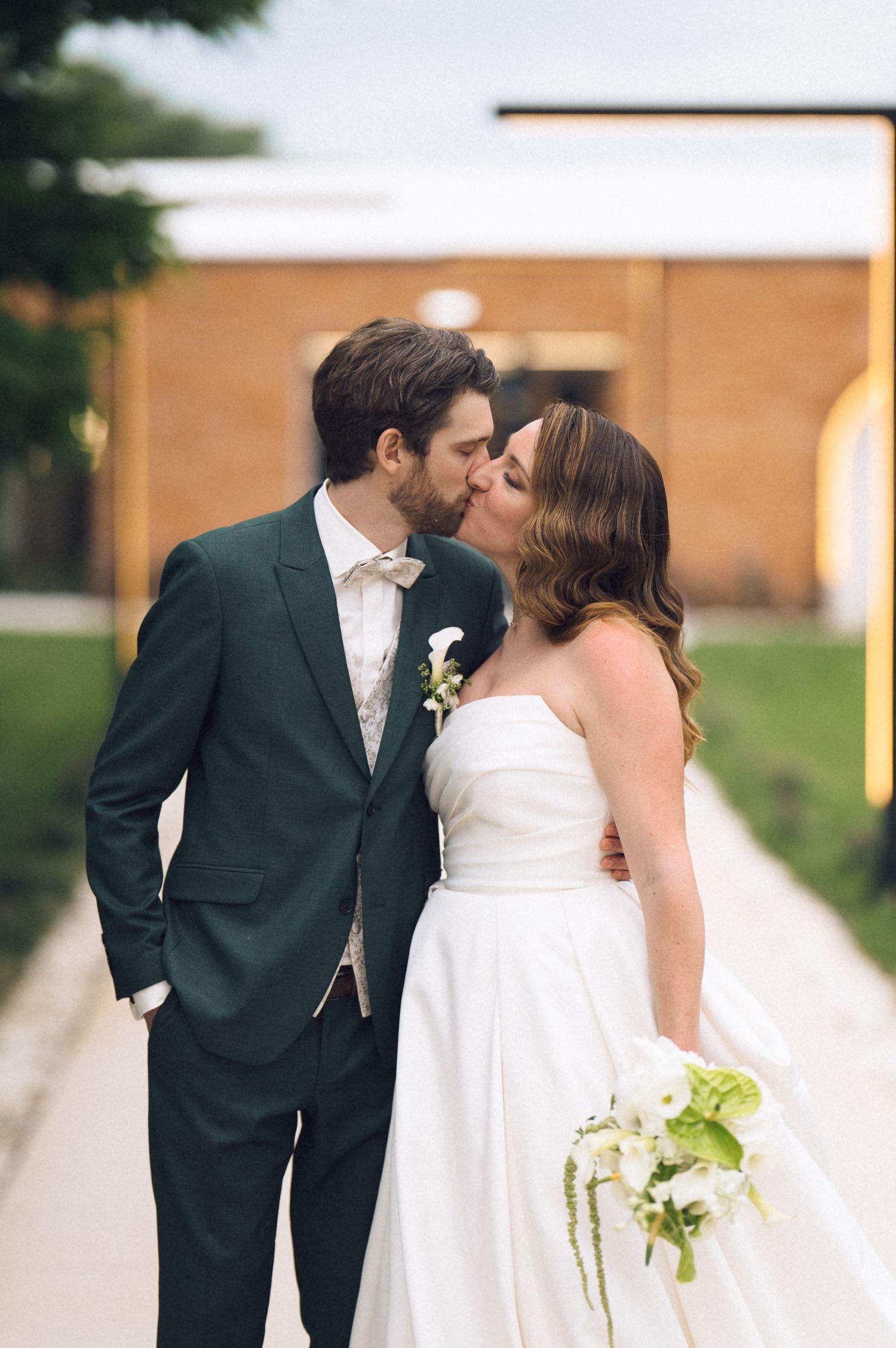 Bride and groom sharing a kiss outdoors, bride in strapless white gown holding green bouquet, groom in dark green suit.