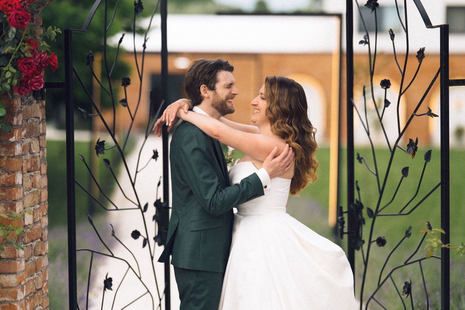 A bride and groom embrace and smile at each other while standing between ornate black iron gates at their wedding venue.