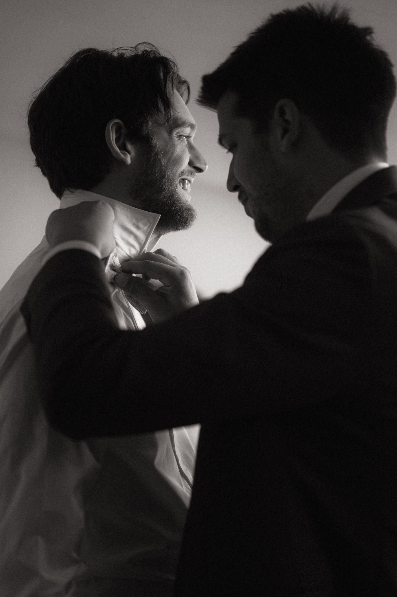 Black and white photo of two men in suits, one helping the other button his shirt collar before a wedding.