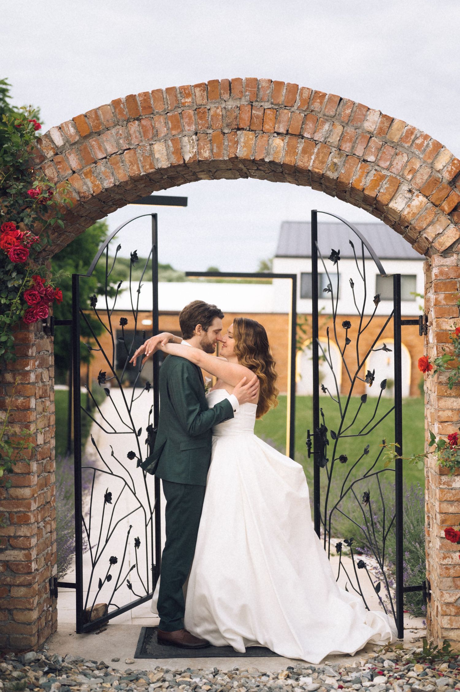 Bride and groom sharing a romantic kiss under a rustic brick arch with ornate iron gates at their wedding venue.