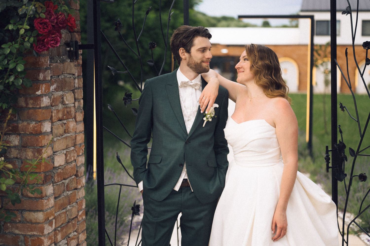 Bride and groom pose together near brick archway, groom in green suit, bride in strapless white gown.