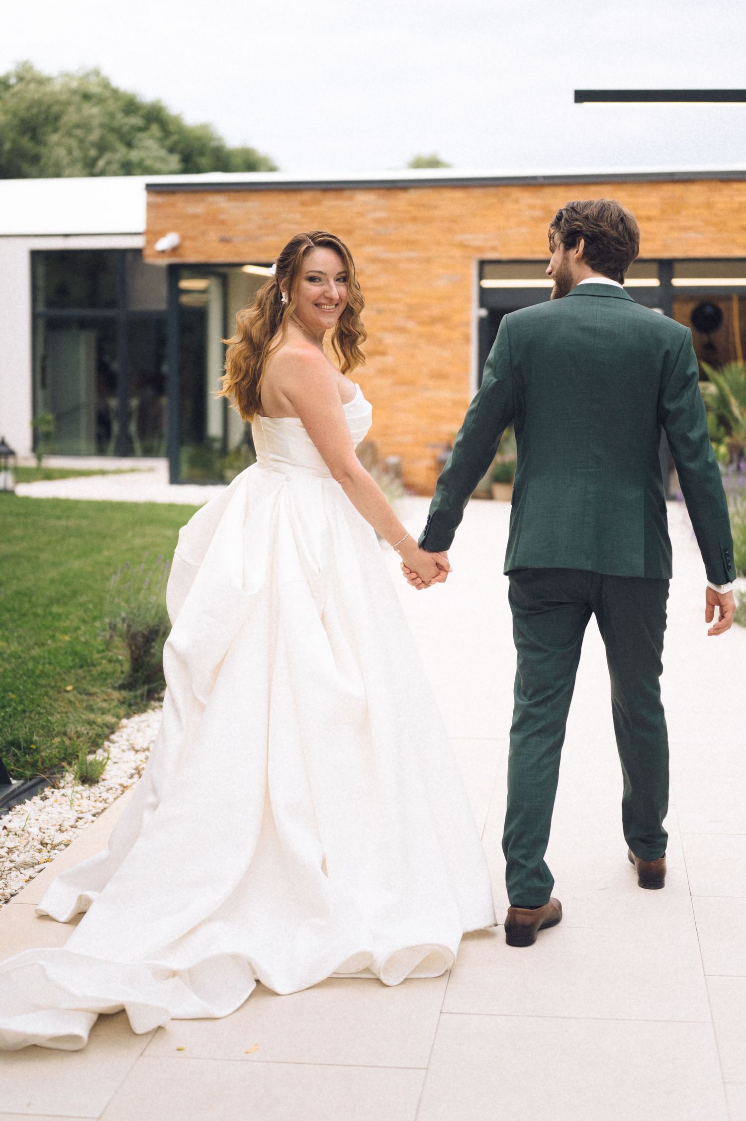 Smiling bride in white ball gown holds hands with groom in green suit walking toward modern building.