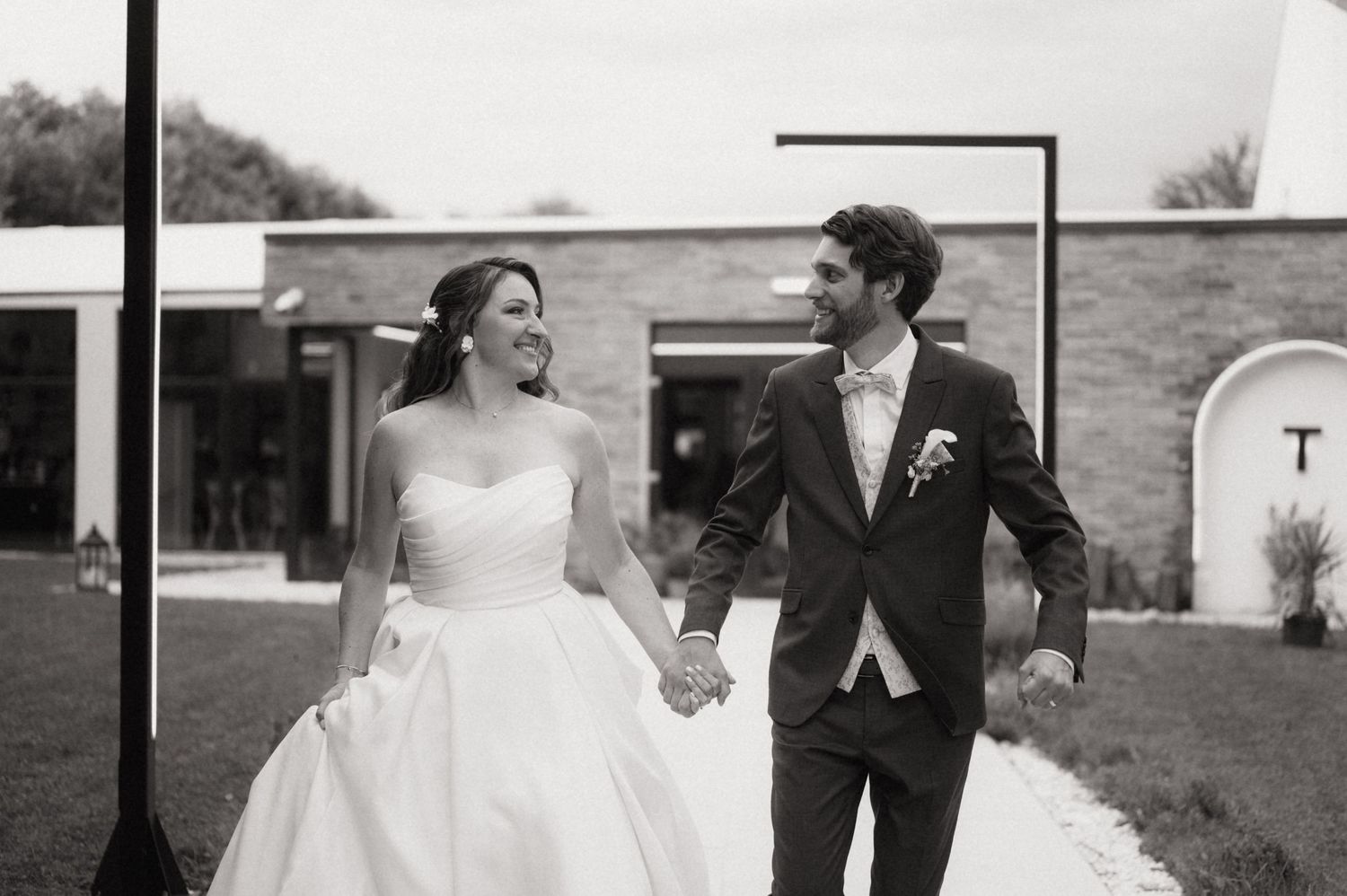 Black and white photo of a smiling bride and groom holding hands walking outside a modern wedding venue.