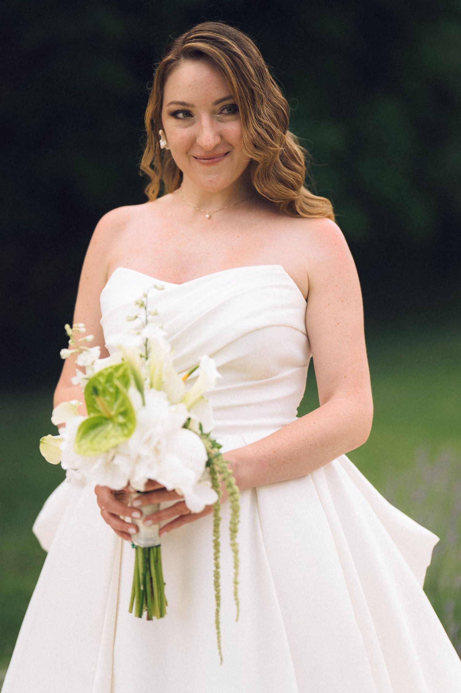 Bride in strapless white gown holding green and white floral bouquet, smiling outdoors with lush greenery background.