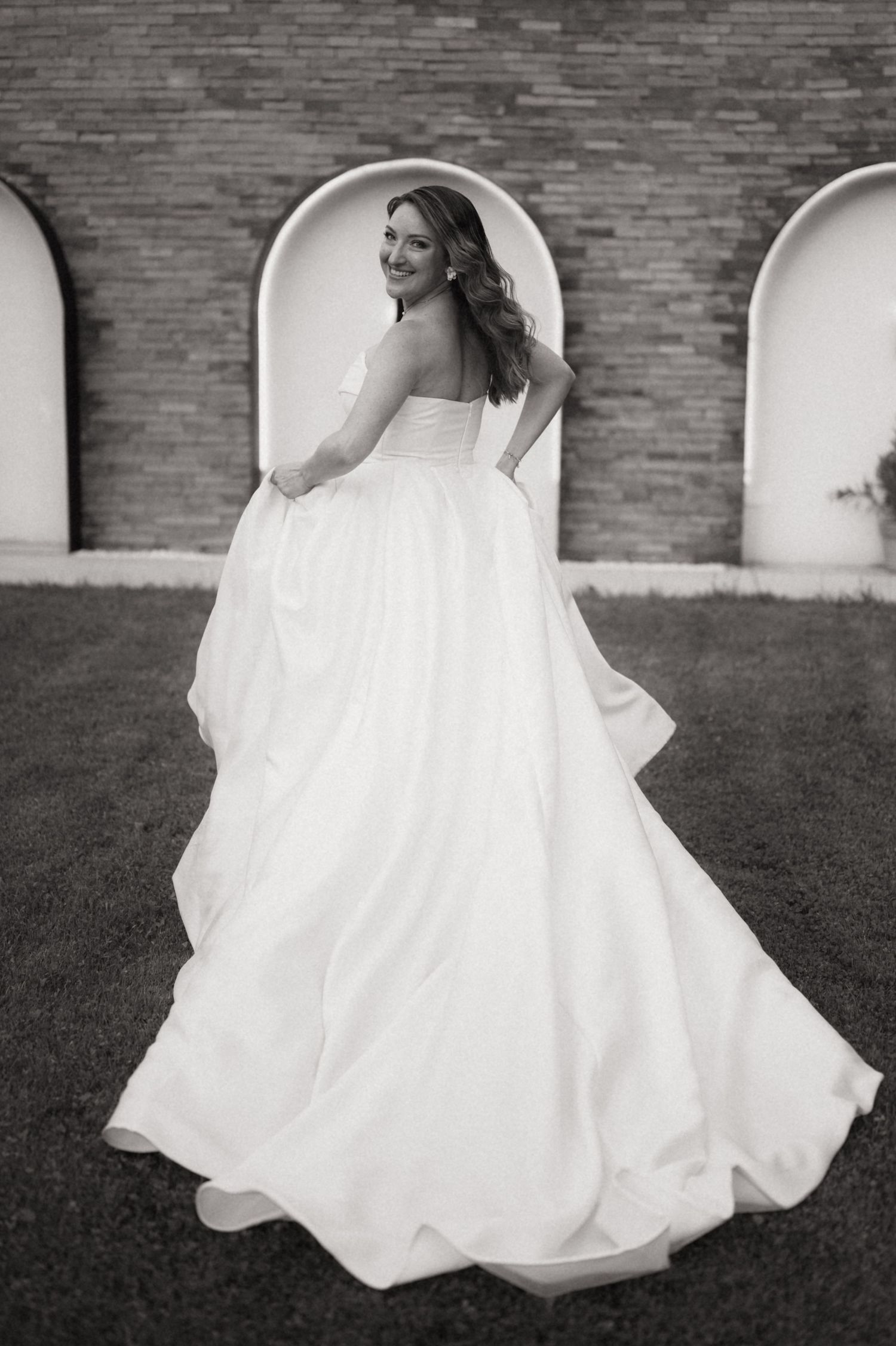 Bride in strapless ball gown wedding dress poses outdoors, glancing back over shoulder near arched brick wall.