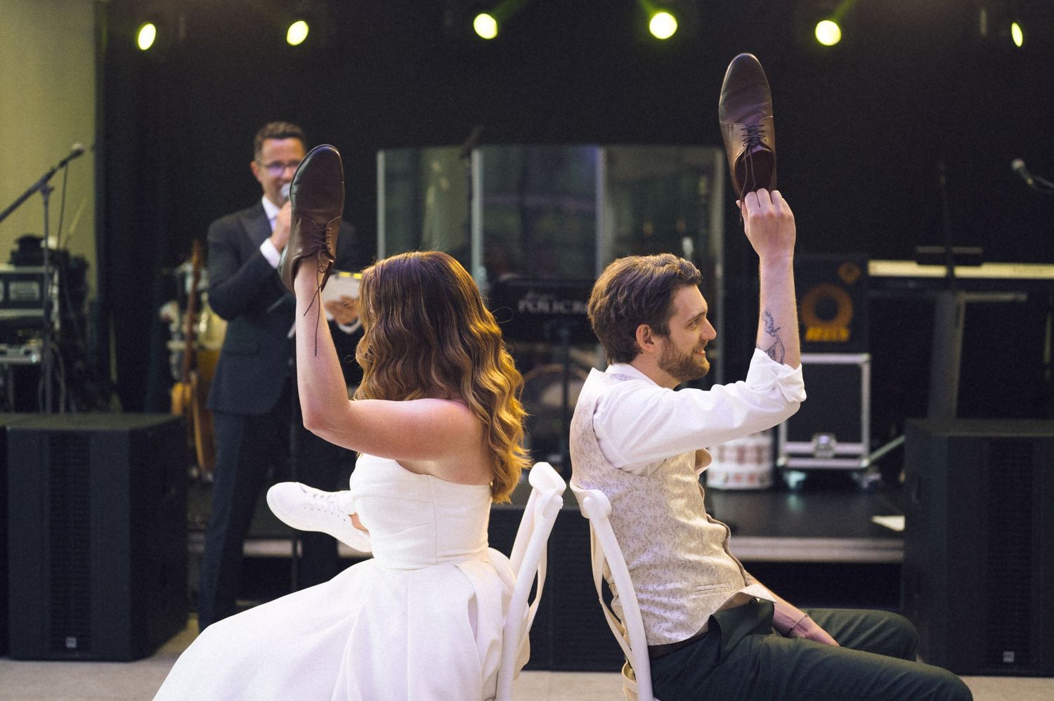 Bride and groom playfully hold shoes up during fun wedding game on dance floor.