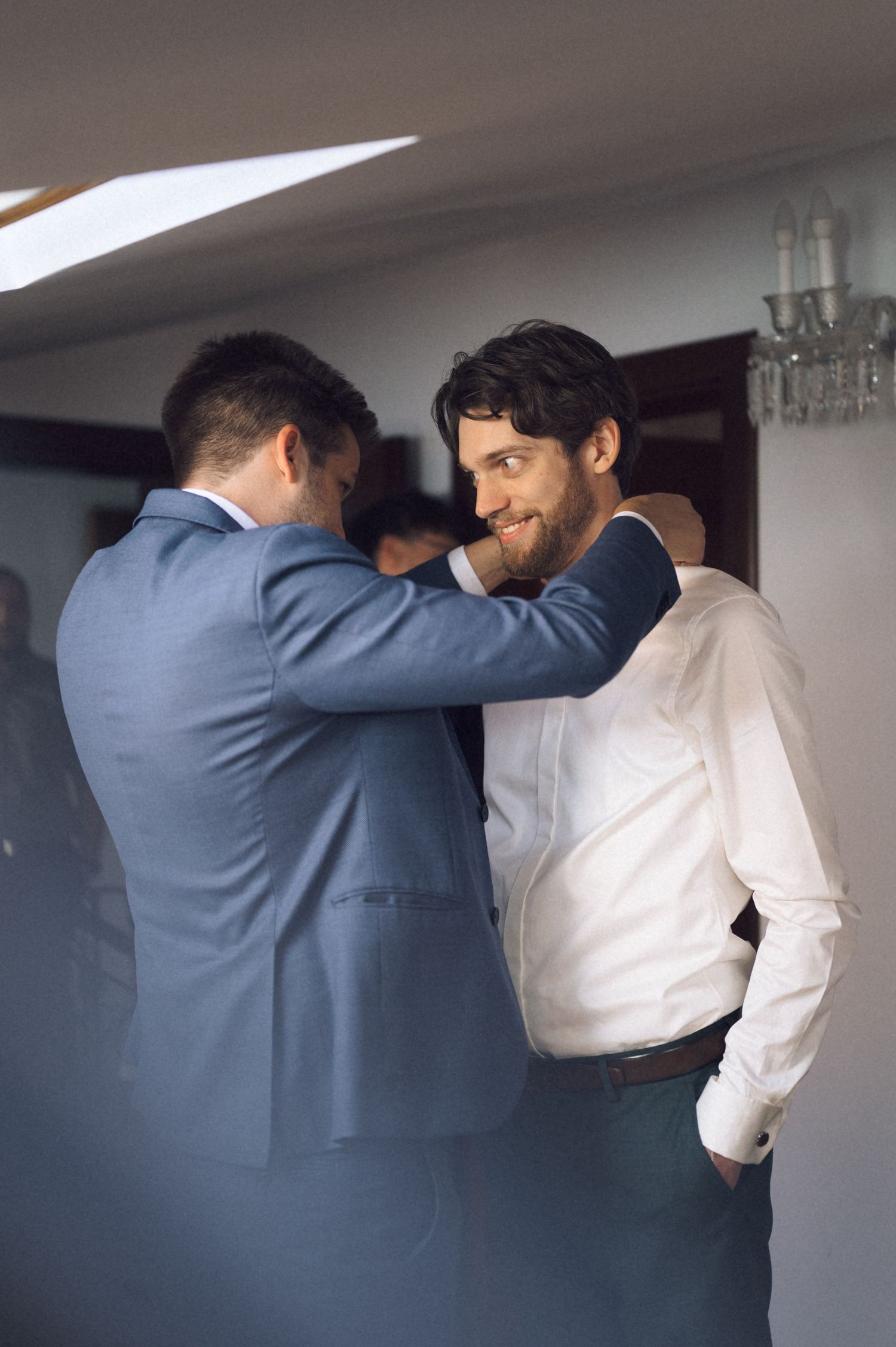 Two men in formal attire help each other prepare, likely groomsmen getting ready for a wedding ceremony.