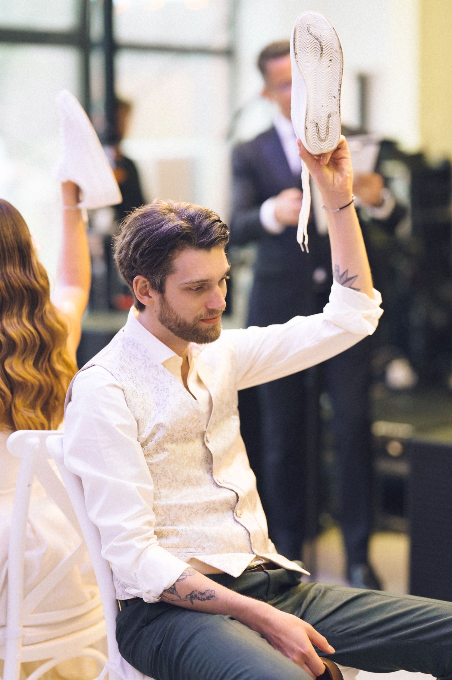 A bearded man in a white vest holds up a sneaker while seated at an indoor event.