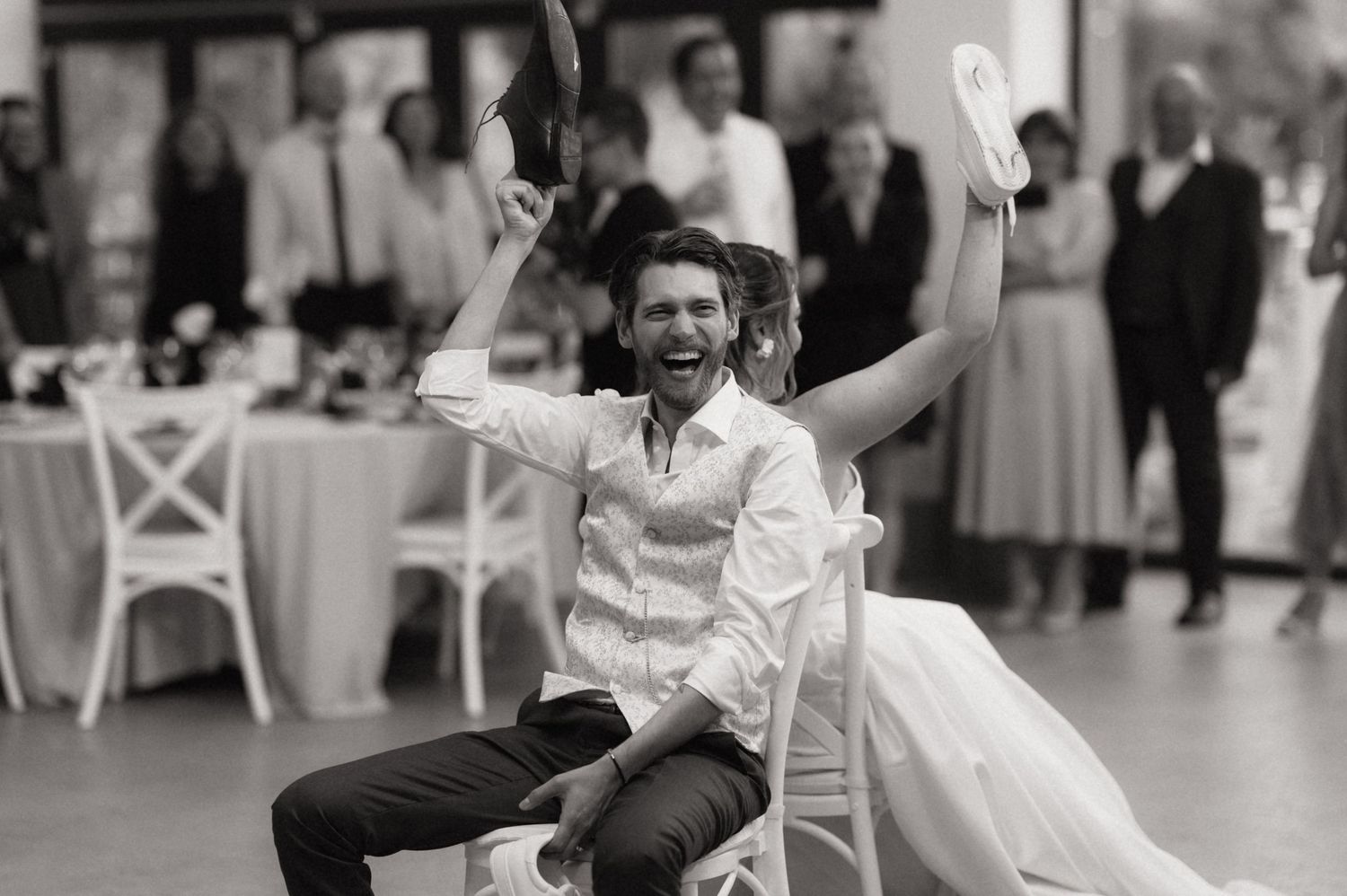 Joyful bride and groom celebrating at wedding reception in black and white photo, both raising arms in excitement.