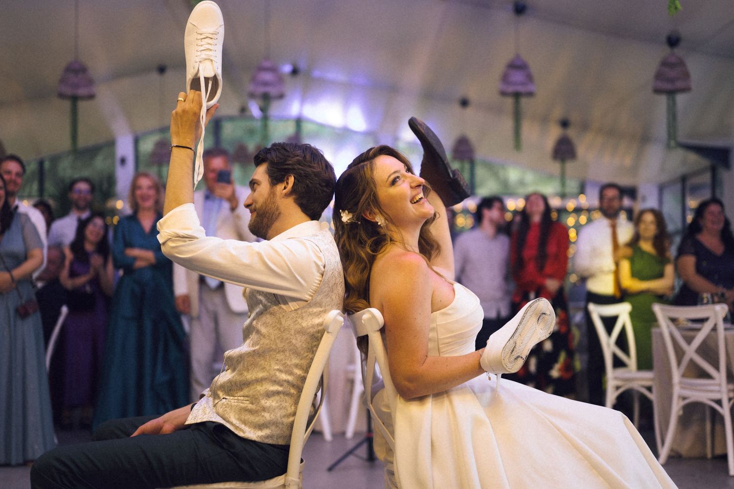 Bride and groom laughing during wedding shoe game at elegant reception, holding up shoes while guests cheer in background.