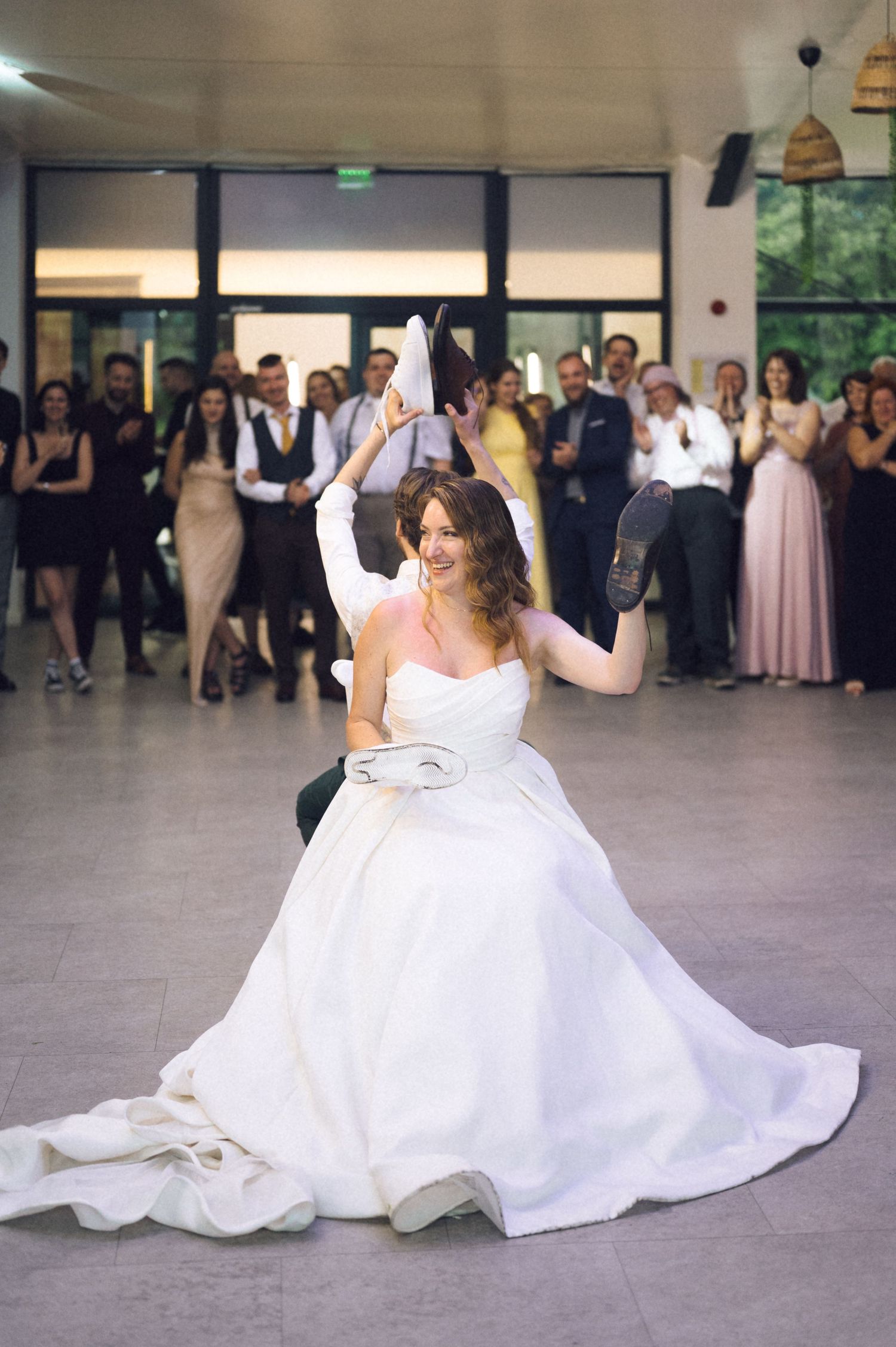 Bride in white ball gown laughing during shoe game at wedding reception with guests watching in background.