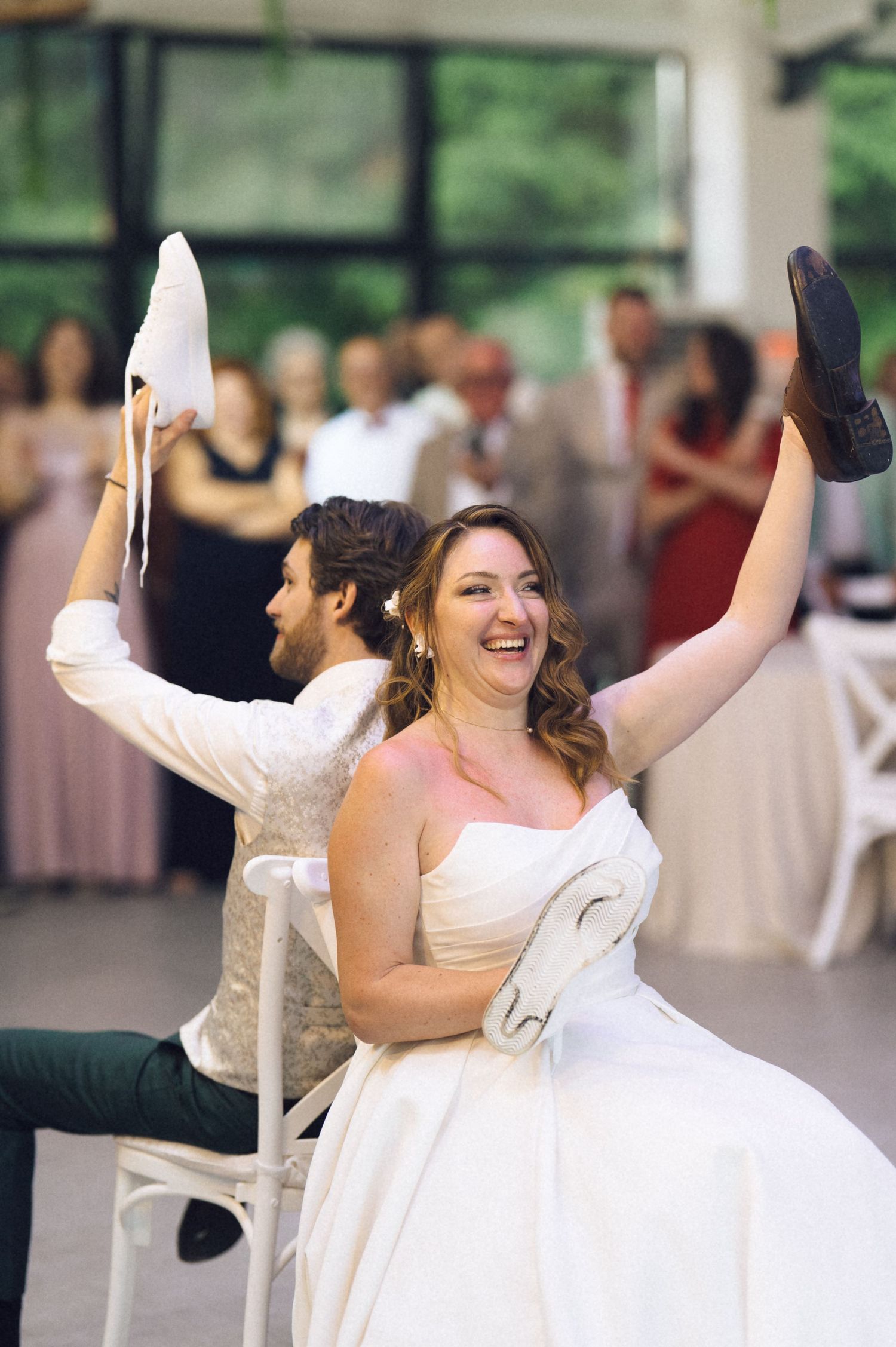 Bride and groom laughing during shoe game at wedding reception, holding up shoes while guests watch and cheer in background.