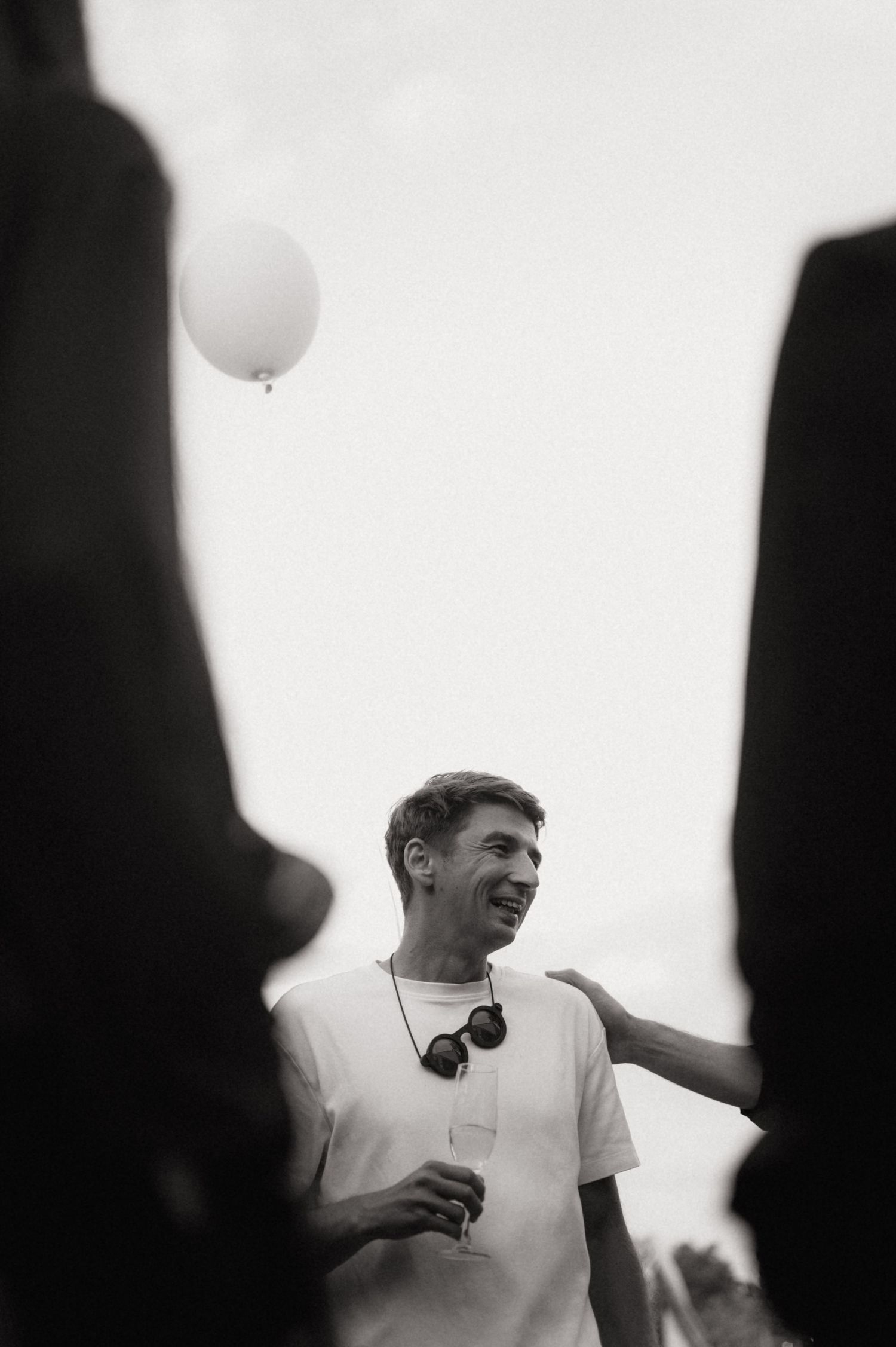 Black and white photo of a smiling man in white t-shirt holding a drink, with a white balloon floating in the background.