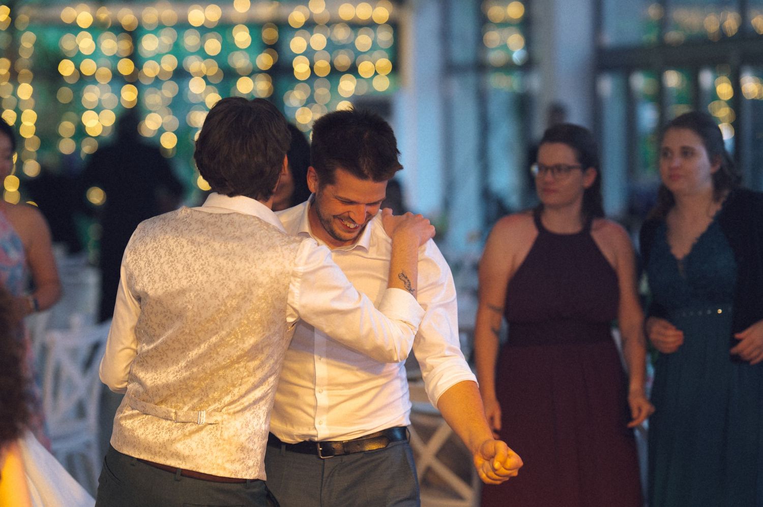 Two men share a warm embrace while dancing at a wedding reception with guests in background.