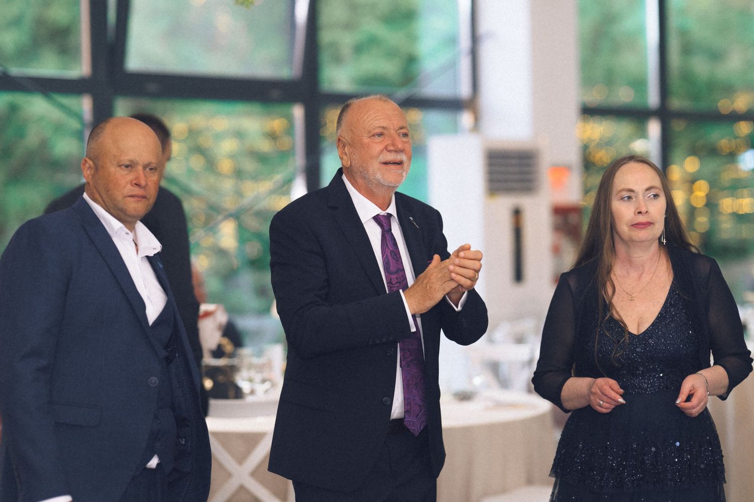 Three elegantly dressed adults socializing at a formal event in a bright venue with lush greenery visible.