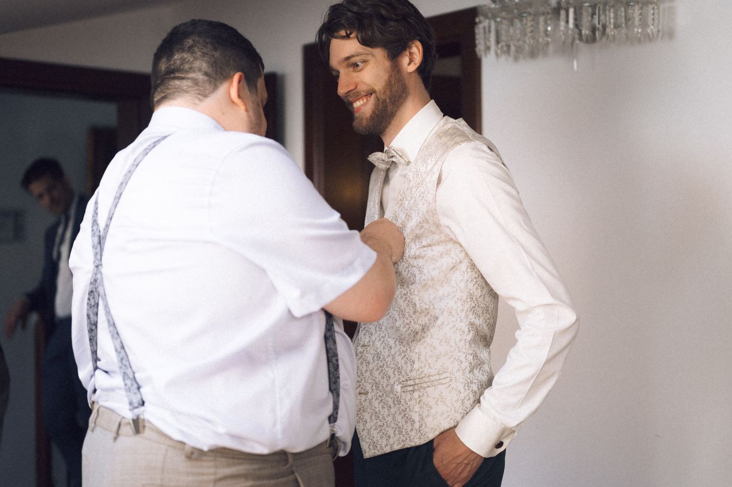 A smiling groom in a floral vest gets his bow tie adjusted by a man in suspenders before a wedding ceremony.