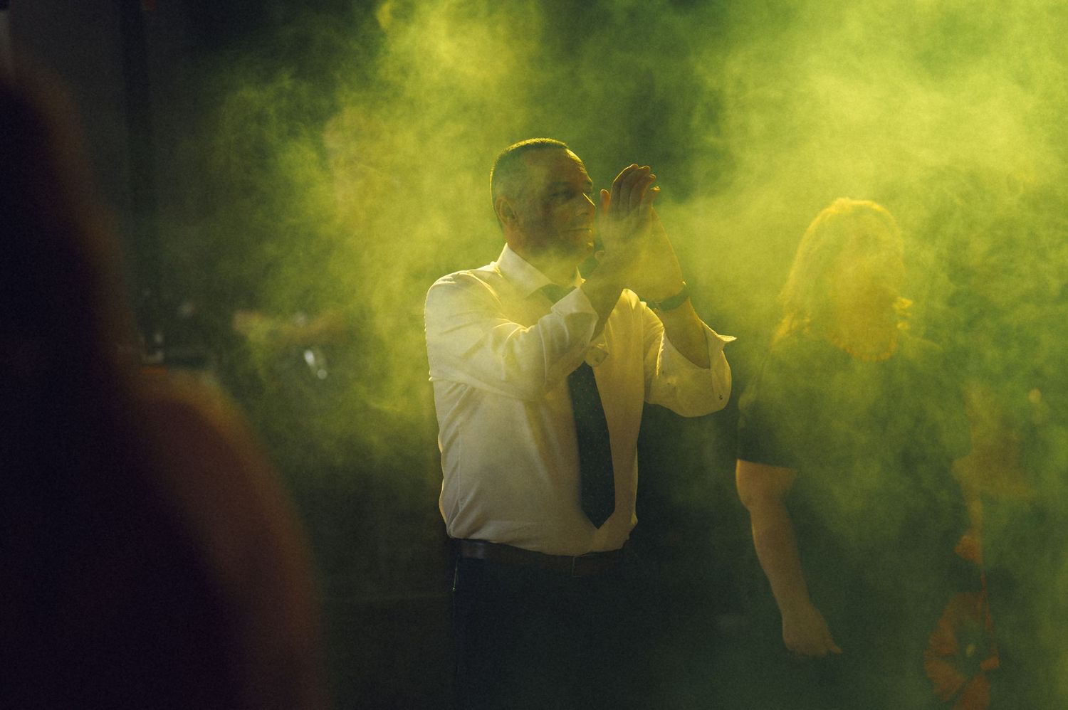 Couple dancing in green smoke at a party, man in white shirt and tie embracing woman in atmospheric fog.