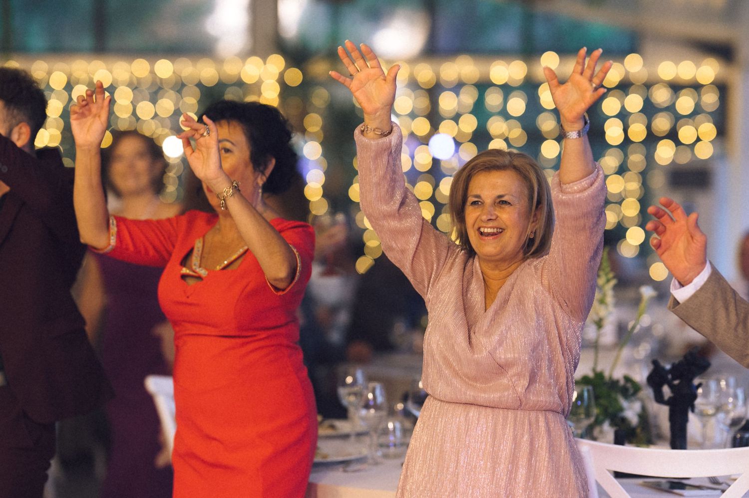 Two women in elegant dresses dancing joyfully with arms raised at a festive wedding reception.