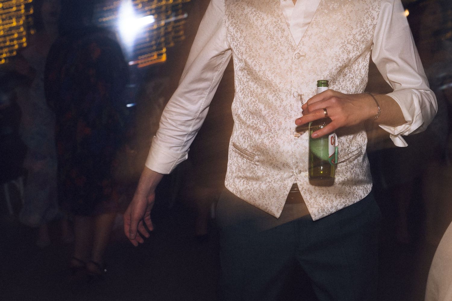 Man in white dress shirt and ornate vest holding a green beer bottle at a formal event or wedding reception.