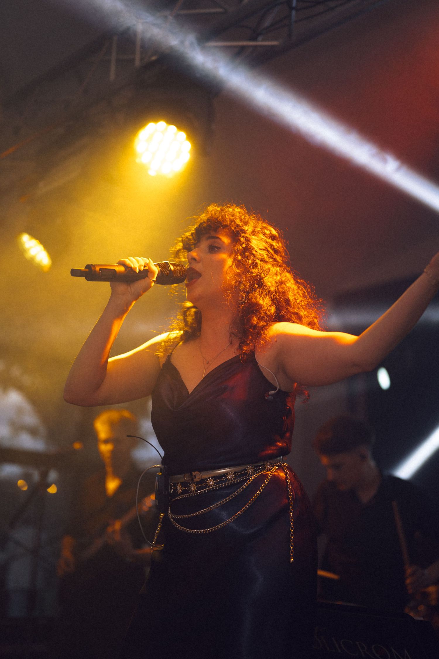 Female singer with curly red hair performs on stage holding microphone, wearing black outfit under dramatic orange stage lighting.