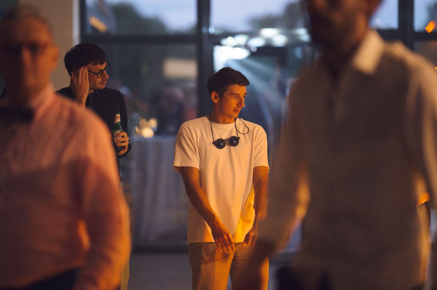 Young man in white t-shirt with sunglasses stands among crowd at evening indoor event.