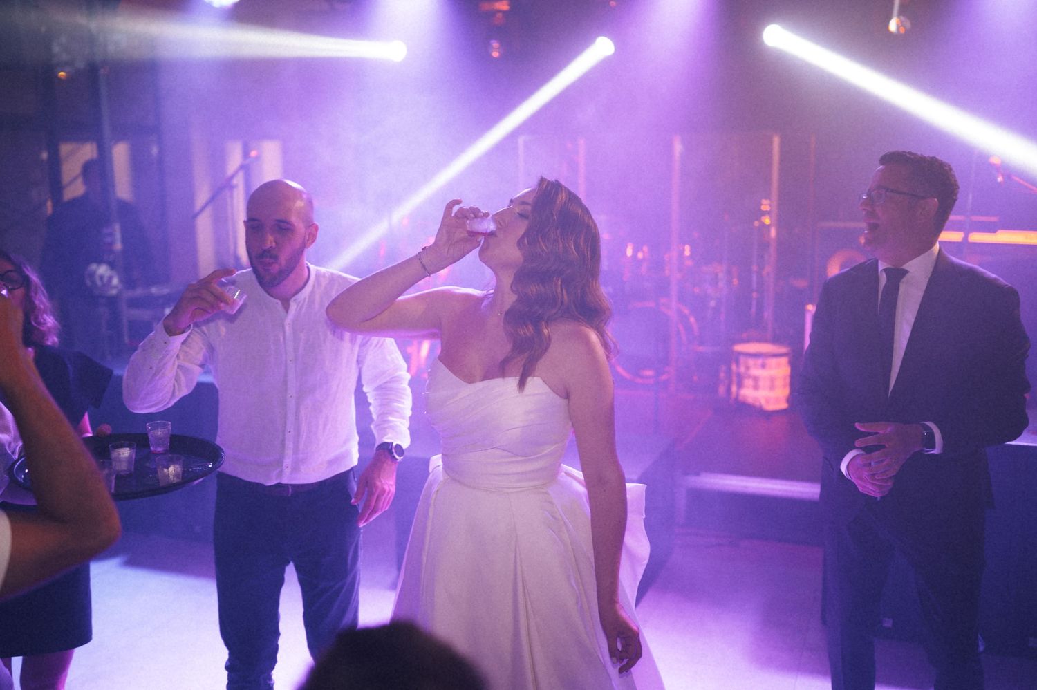 Bride and groom celebrating on a purple-lit wedding dance floor, groom drinking a shot while guests watch nearby.