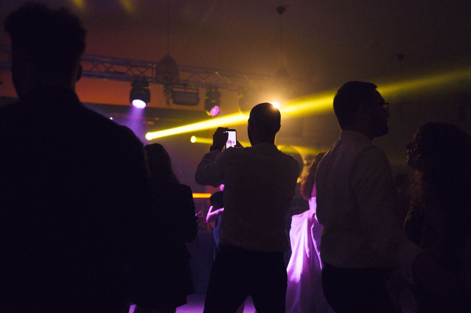 People dancing at a nightclub with colorful purple and yellow stage lights illuminating the dark venue.