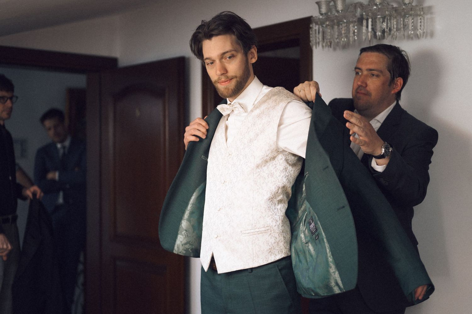 A man being helped into a green suit jacket by another man in a formal indoor setting.