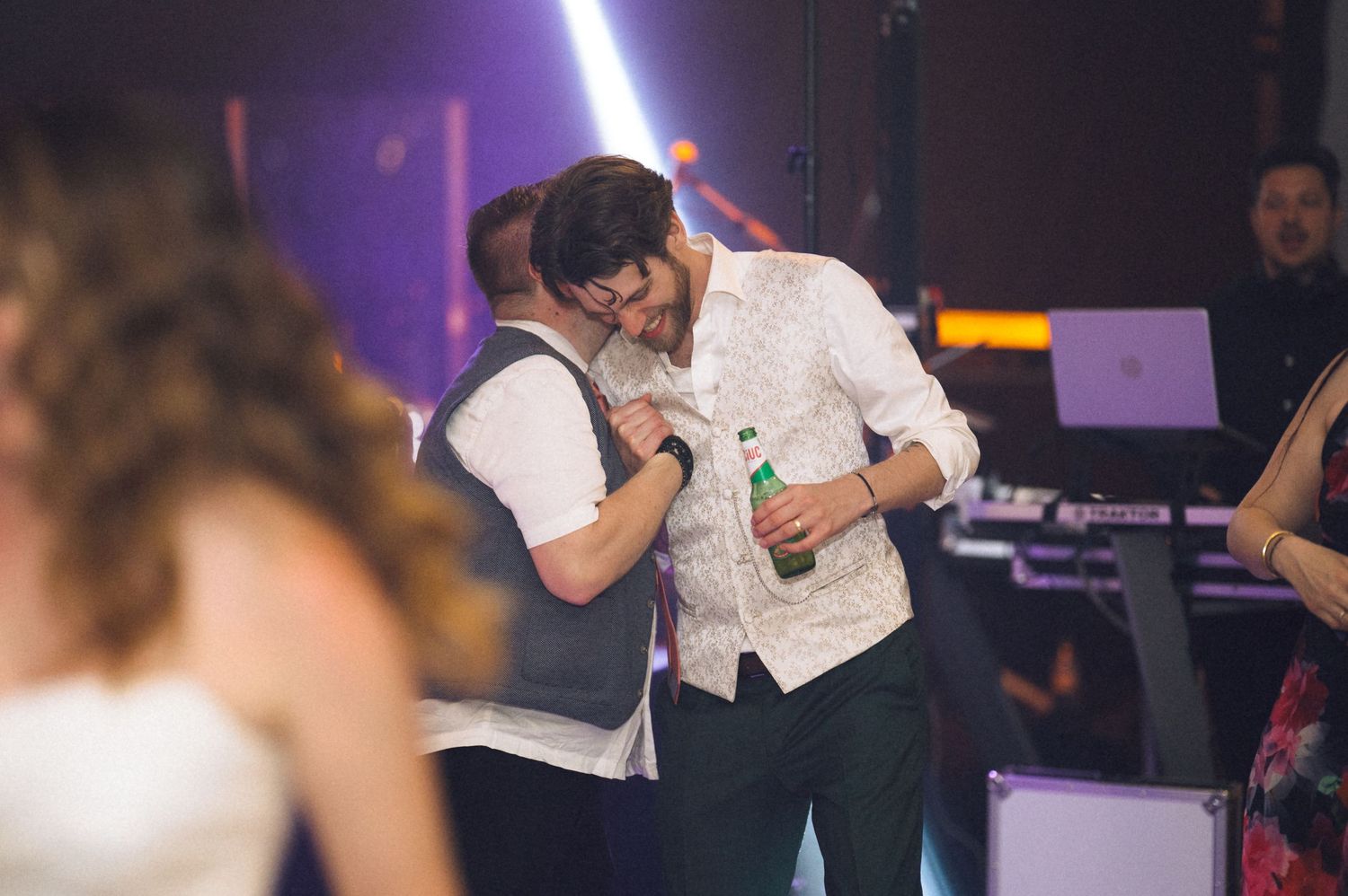 Two men dancing and laughing together at a lively wedding reception party with stage lights in background.