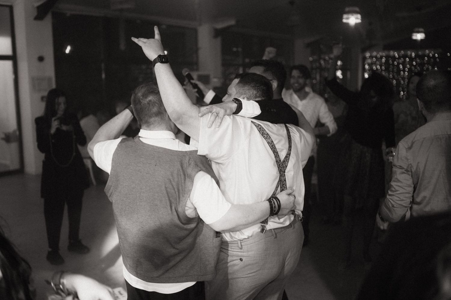 Black and white photo of guests dancing and embracing at a lively indoor party or wedding reception.